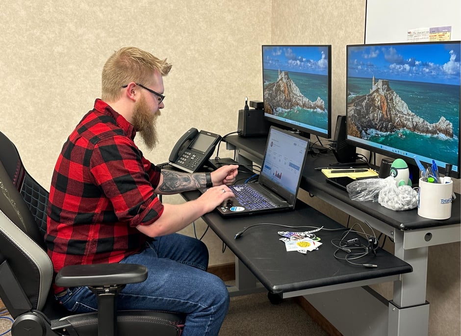 a man in a flannel shirt working at a desk with 2 monitors